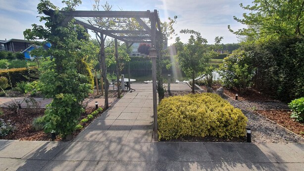 Un sendero en un jardín con un pérgola de madera, rodeado de arbustos y árboles diversos.