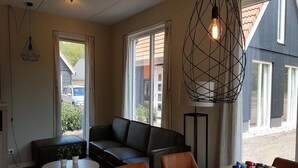 Una sala de estar moderna con un sofá de cuero negro, una mesa redonda y una mesita de centro. Hay dos ventanas con cortinas, una lámpara colgante y vistas a un edificio.