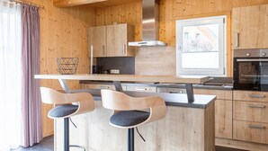 A modern kitchen with wooden cabinetry, a stainless steel range hood, and a built-in oven. There are two bar stools with cushioned seats. - Luxurious Chalet in Murau with Outside Hot Tub (Murau)
