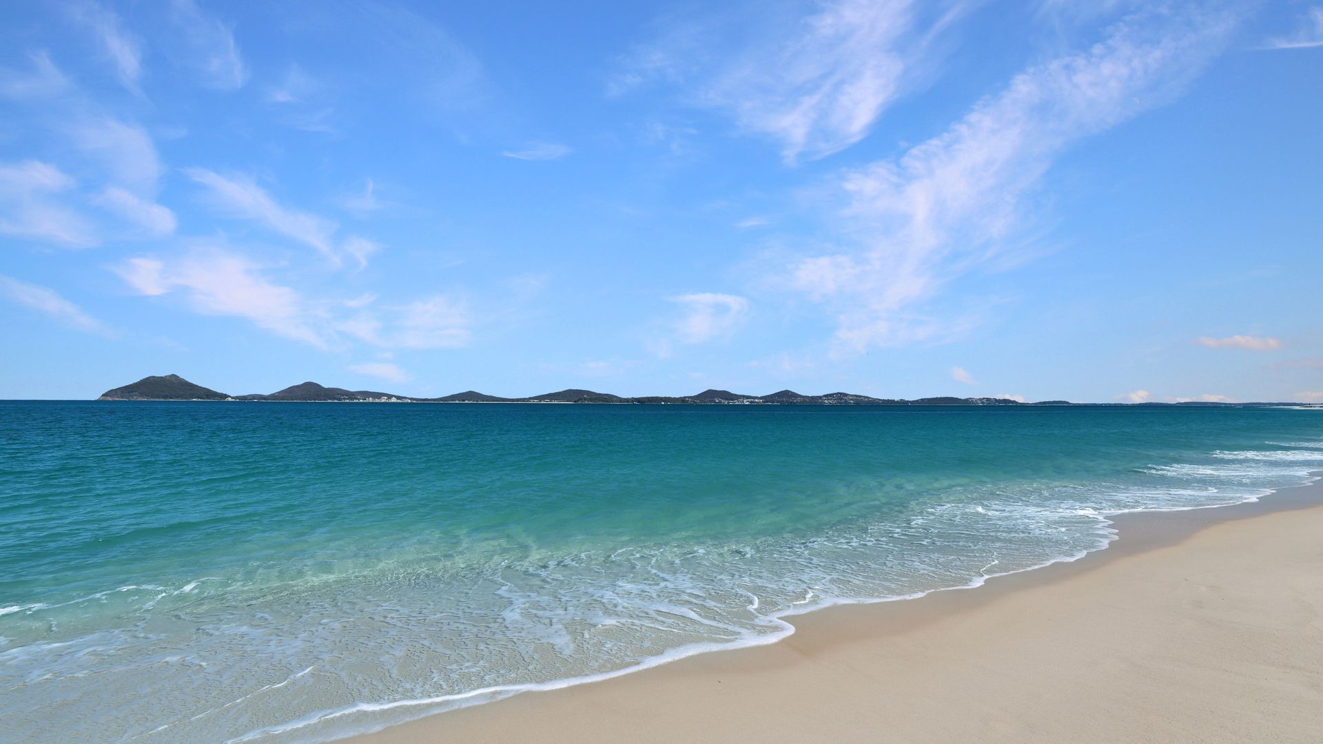 Una playa con aguas turquesas cristalinas, una orilla arenosa y colinas al fondo.