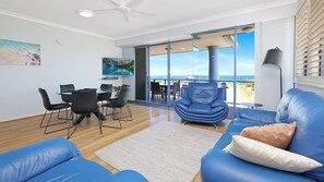 Un salon moderne avec un canapé bleu, un coin repas avec des chaises et une grande fenêtre donnant sur la mer.