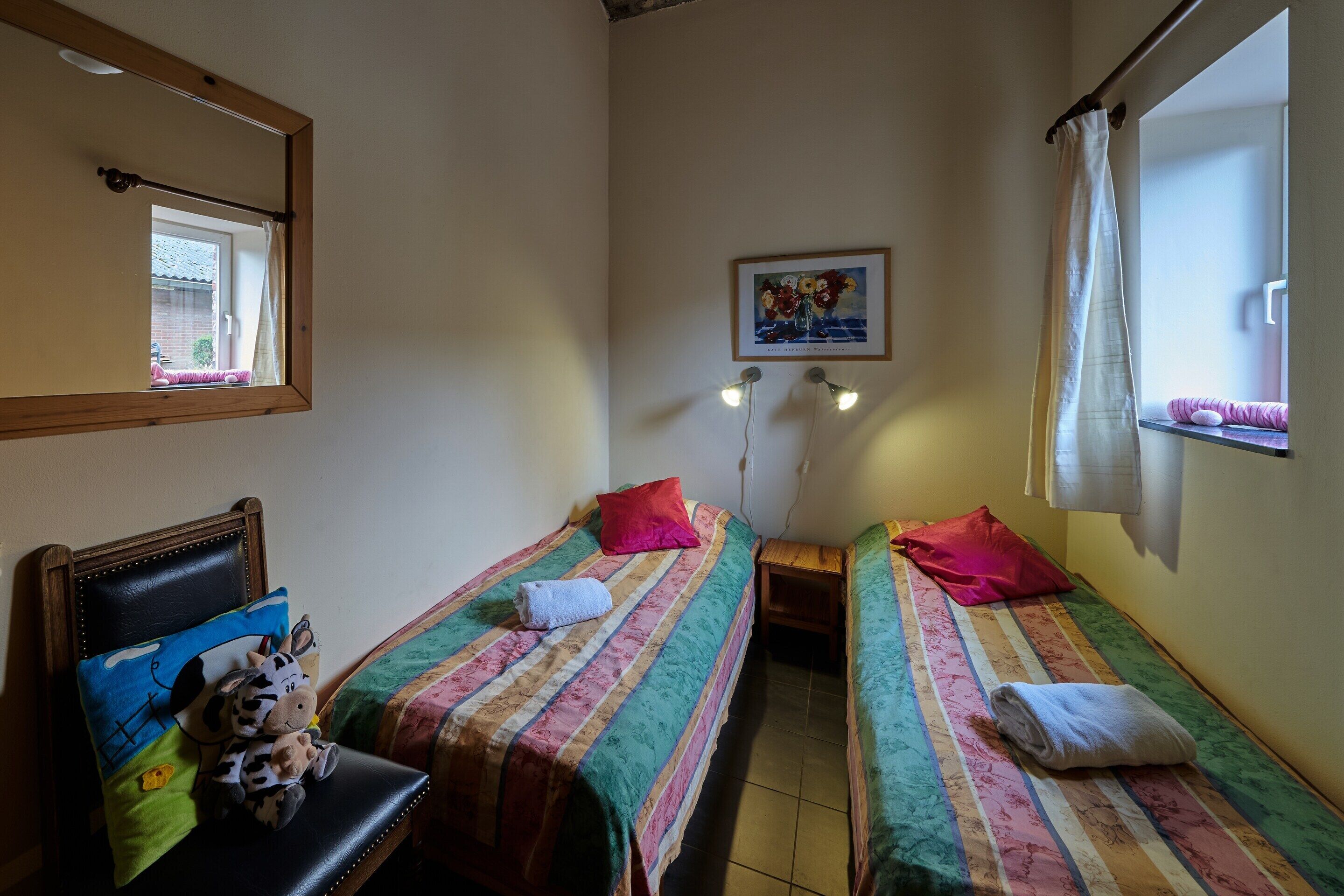 Une chambre avec deux lits, une petite table, une chaise, un miroir, une fenêtre avec un rideau et un tableau accroché au mur.