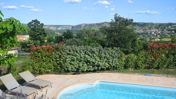 Ein Schwimmbecken mit Liegestühlen, umgeben von Grün und Blick auf eine Stadt in der Ferne.