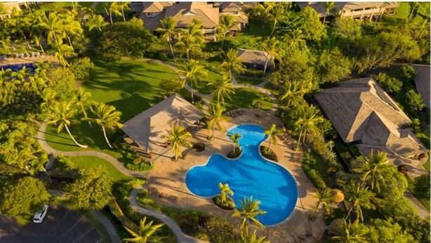 Eine Vogelperspektive zeigt einen luxuriösen Urlaubsort mit einem zentral gelegenen Schwimmbad, umgeben von Palmen und Hotelgebäuden.