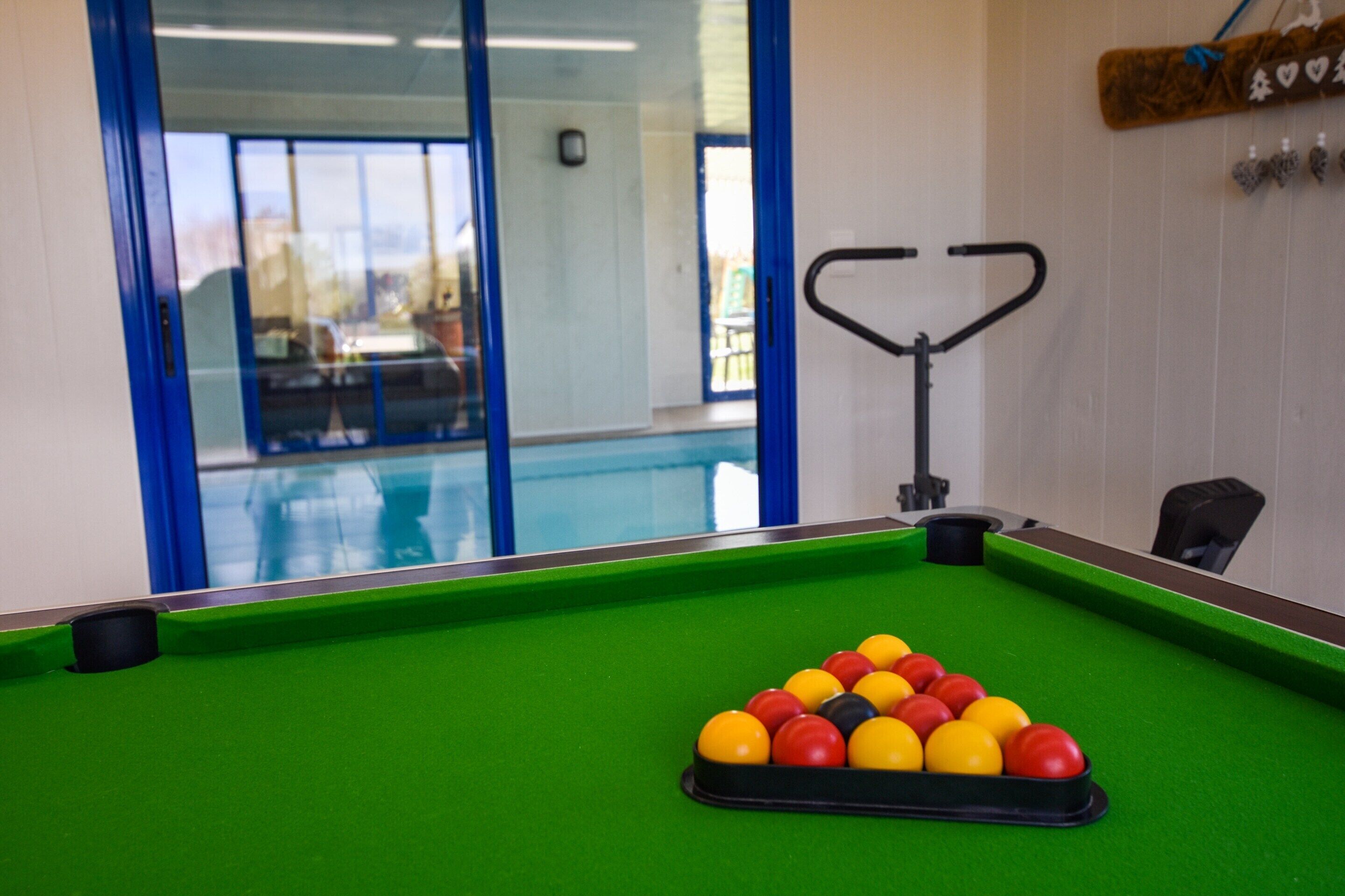 Une table de billard avec les billes disposées en forme de triangle, un queue de billard et une fenêtre donnant sur une piscine.