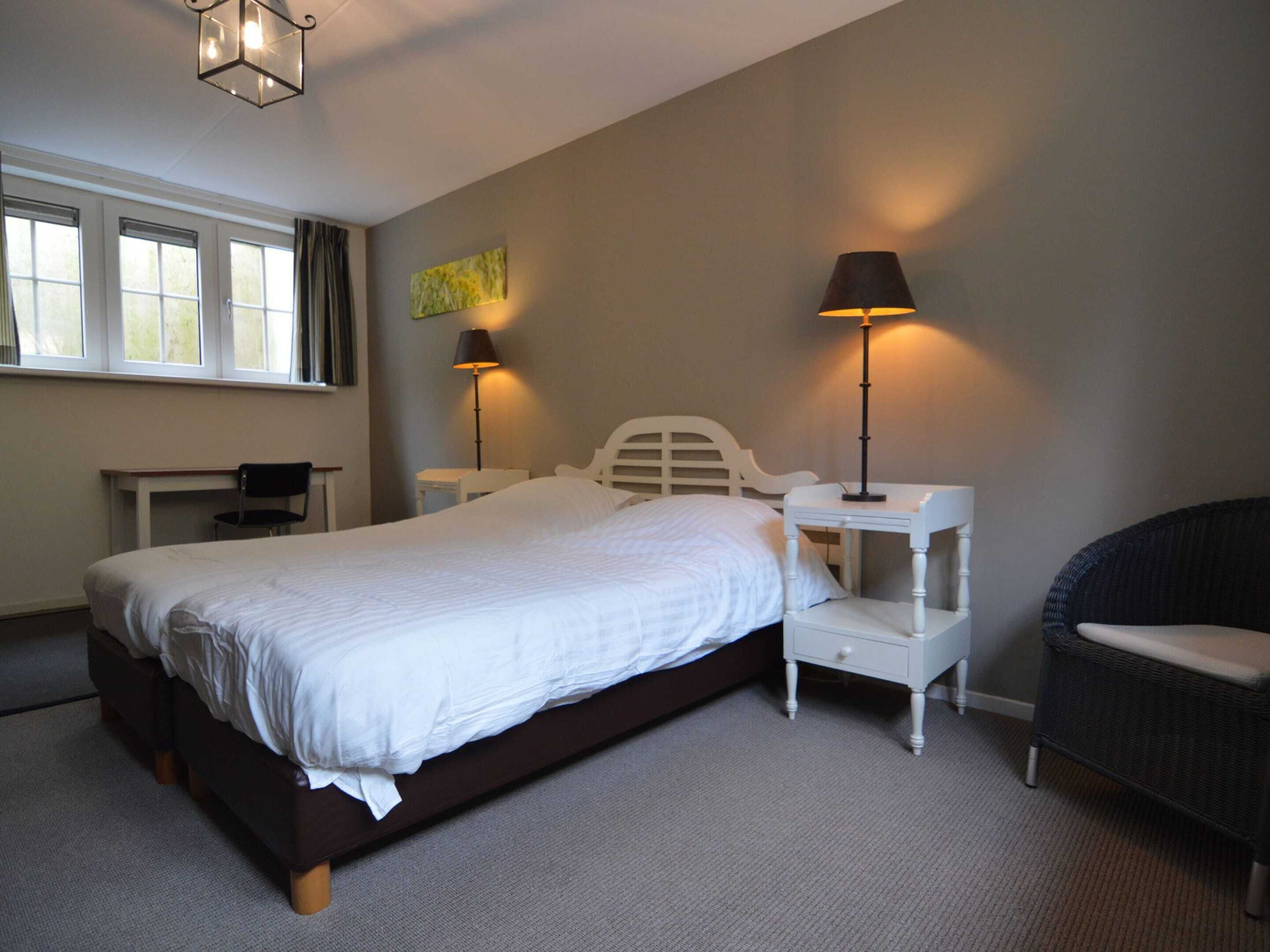 A bedroom with a large bed, two bedside tables with lamps, a chair, a desk, and a window with curtains.