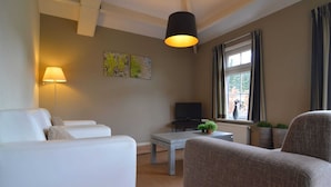 Un salon moderne avec un canapé blanc, un fauteuil marron, une table basse, un téléviseur à écran plat et une grande fenêtre avec des rideaux.