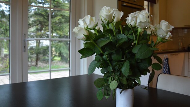 Un vase blanc contenant des roses blanches, posé sur une table sombre dans une pièce dotée d’une fenêtre, avec une cuisine en arrière-plan.