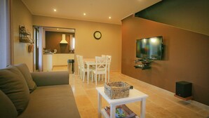 Een woonkamer met een bank, een eettafel met stoelen, een televisietoestel en een klok aan de muur.
