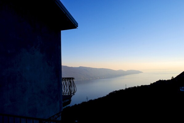 Un balcon donnant sur un lac paisible, avec des montagnes lointaines à l’horizon, un ciel dégagé et un bâtiment à la façade sombre.