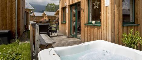 A wooden cabin with a hot tub, outdoor seating, and a view of other cabins in the background.