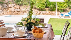 Une table à manger dressée avec des assiettes, des bols, une théière et un panier de pain, avec une piscine et de la verdure en arrière-plan.