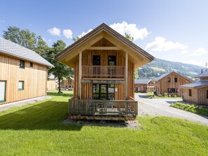 En trästuga med en balkong, omgiven av andra liknande byggnader, belägen på en gräsyta med ett berg i bakgrunden.