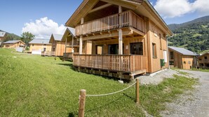 A wooden chalet with a balcony, surrounded by a grassy area and other similar buildings, set against a mountainous backdrop.