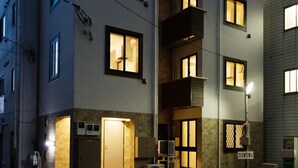 A multi-story residential building with illuminated windows and balconies, featuring a ground-level entrance with a door and a small outdoor area with a trash bin. - Ginza Tsukiji GUEST HOUSE 301. (Chuo)