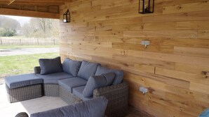 A covered outdoor seating area with a wicker sectional sofa and ottoman, wooden walls, and two wall-mounted lanterns. - From the Zealand Coast and Near Brugge (Aardenburg)