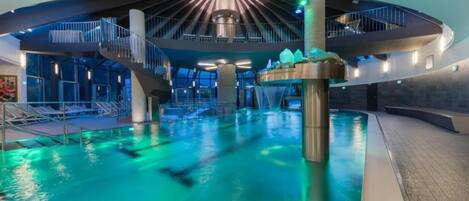 A modern indoor swimming pool with a central water feature, surrounded by a circular viewing area with glass walls.