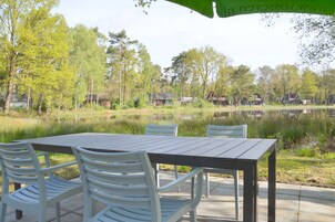 A wooden table with chairs set outdoors, overlooking a lake with houses in the background.