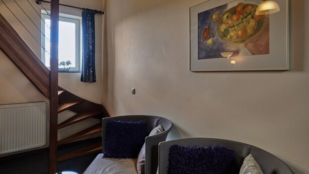 Una habitación con una escalera, una cama con almohadones azul oscuro, un cuadro enmarcado de comida y una ventana con cortinas.