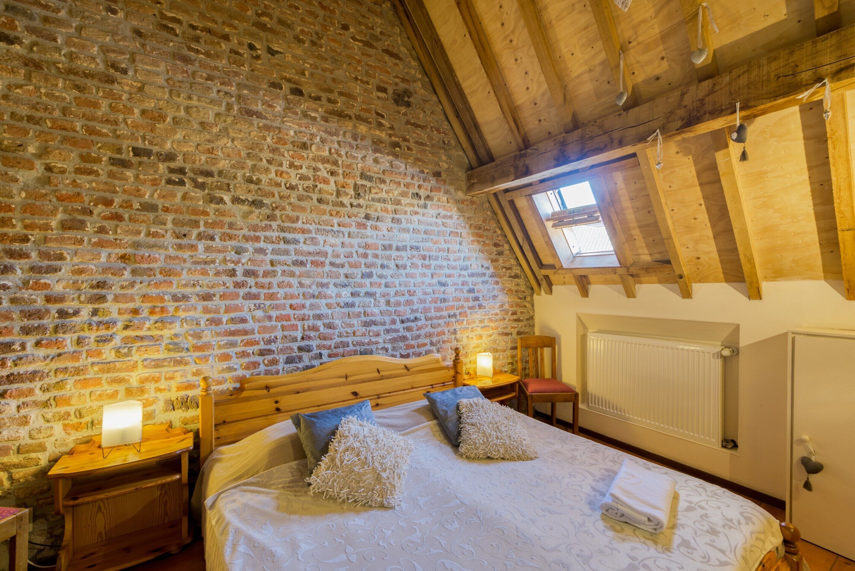 Une pièce chaleureuse avec un mur en briques, des poutres en bois au plafond, un lit avec des oreillers, une table de chevet avec une lampe et une fenêtre donnant sur un beau paysage.