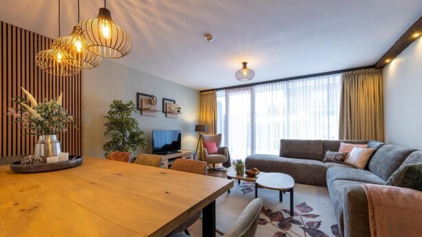 Een moderne woonkamer met een houten eettafel, een bank en een televisie. Er staan planten en decoratieve objecten aan de wanden.
