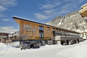 Un bâtiment moderne avec une façade en bois et une enseigne sur laquelle on peut lire « ACHSER KURZEN ». Des voitures sont garées devant, et des montagnes enneigées se dressent à l'arrière-plan.