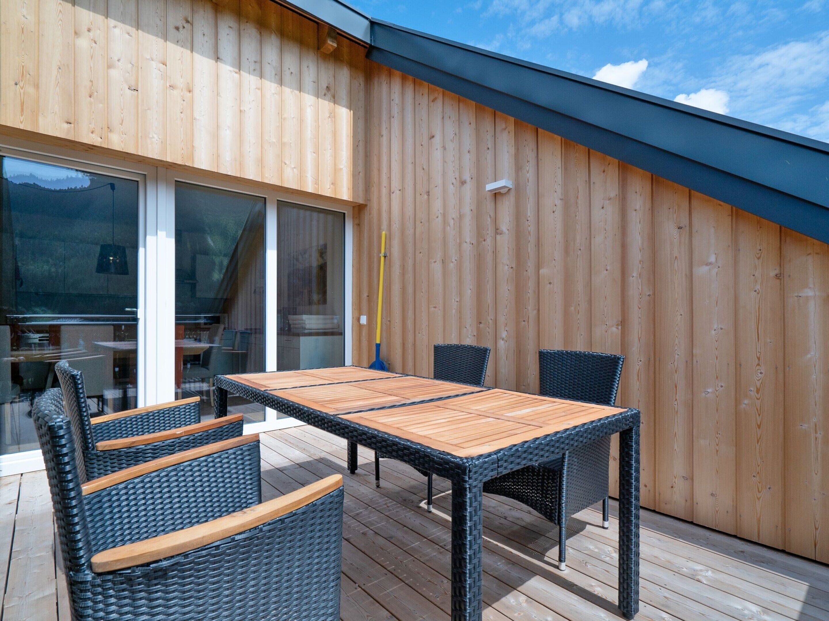 An outdoor deck with a wooden table and wicker chairs, a glass door leading inside, and a wooden wall.