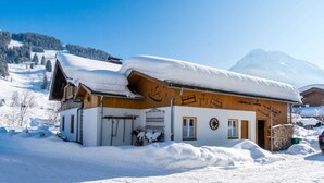 Un chalet de madera cubierto de nieve con techo inclinado, un porche de madera y montañas al fondo.