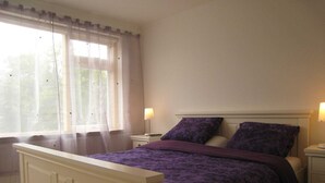 Ein Schlafzimmer mit Bett, Nachttisch, Fenster mit Vorhängen und einer Lampe.