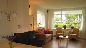 Ein Wohnzimmer mit Esstisch aus Holz, einer Couch, zwei Sesseln, einer Stehlampe und einem Fenster mit Ausblick.