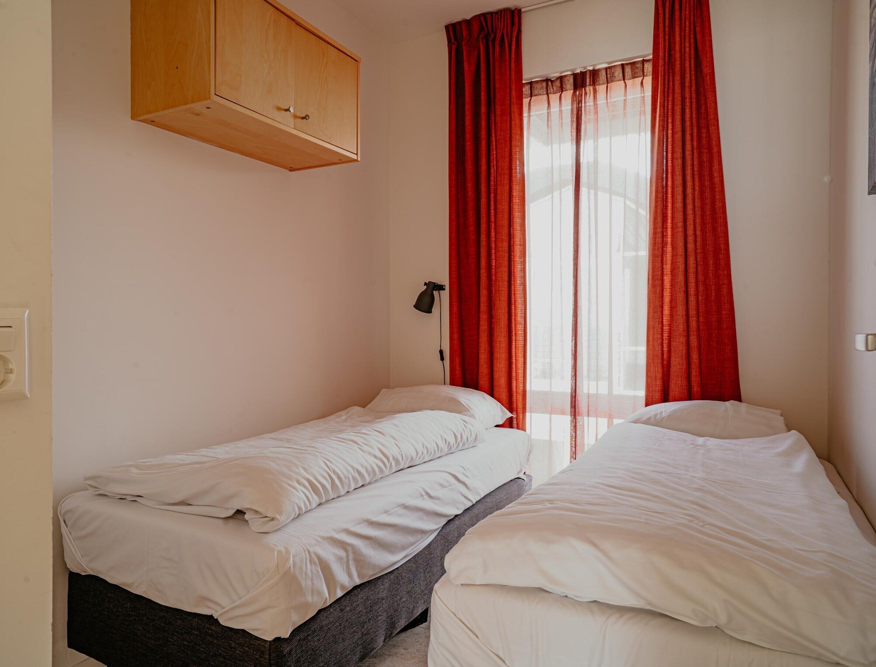 Ein kleines Schlafzimmer mit zwei Einzelbetten, einem Holzschrank über dem Bett links und roten Vorhängen am Fenster.