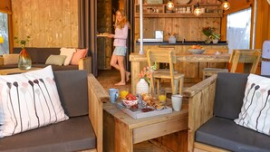 A cozy cabin interior with a wooden dining area, a sofa, and a breakfast tray with fruits and pastries. - Campsite - Combined Tents With Kitchen and Bathroom Located Near a Pond (Bant)