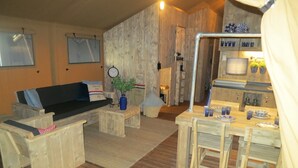 A rustic cabin interior with a wooden floor, a sofa, a dining table with chairs, a small kitchen area, and a clock on the wall. - Campsite - Combined Tents With Kitchen and Bathroom Located Near a Pond (Bant)