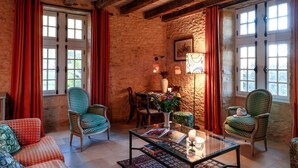 A cozy room with stone walls, wooden beams, and red curtains. There are two green patterned chairs, a glass coffee table with books, and a small table with a vase and candles. - Retreat Near Siorac-en-perigord (Saint-Germain-de-Belvès)