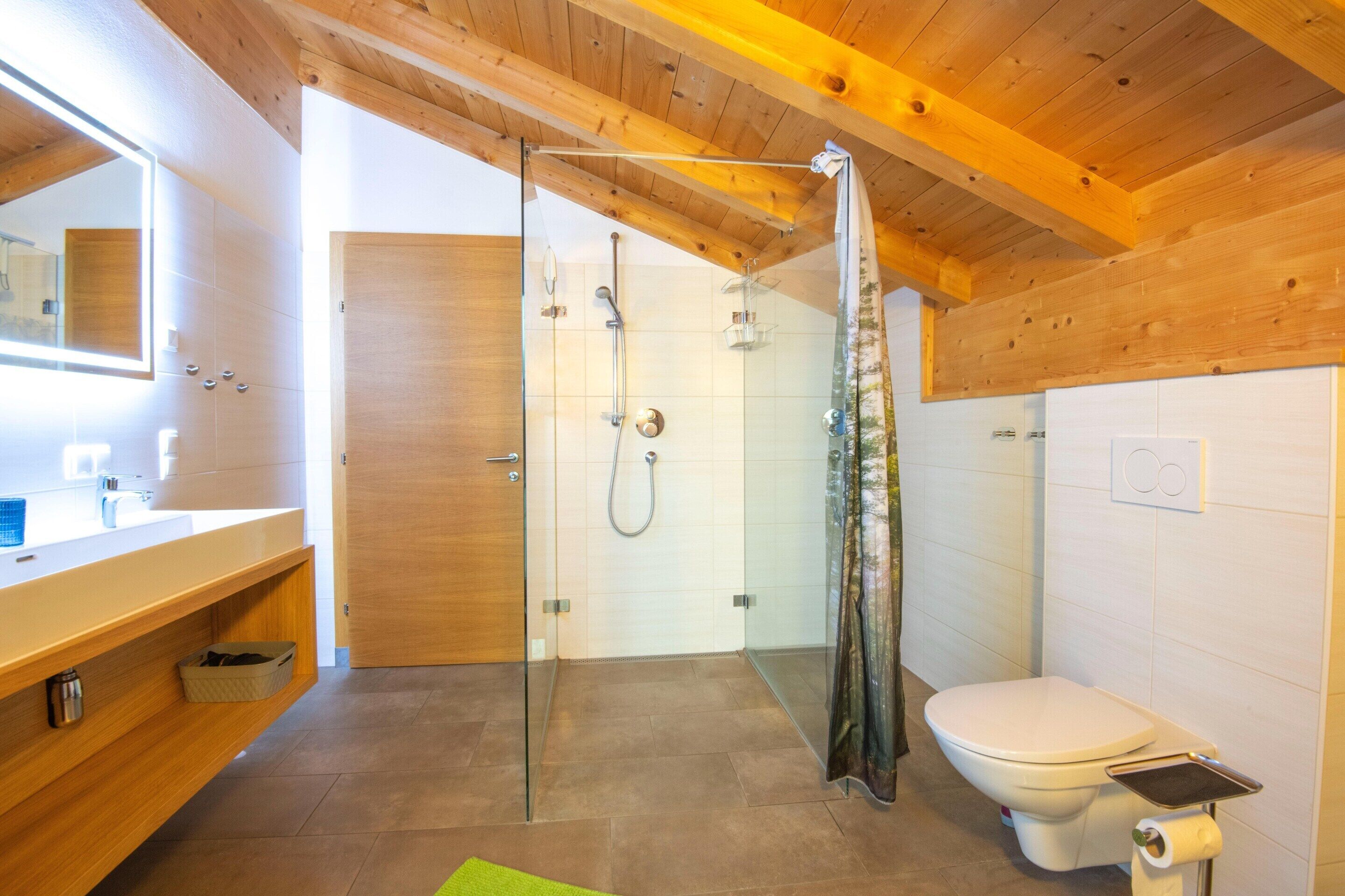 Une salle de bain moderne dotée d’une douche à parois de verre, de toilettes avec un porte-rouleau de papier hygiénique et d’un meuble-lavabo en bois avec un miroir.