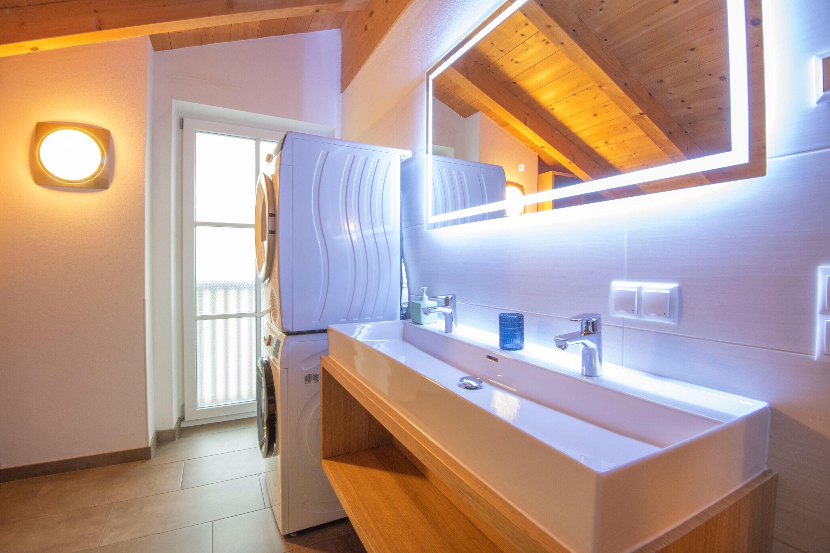 Een moderne badkamer met een wit wastafelmeubel, spiegel en houten kast. Een wasmachine en handdoekrek zijn zichtbaar.