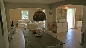 A dining area with a wooden table and chairs, a hanging light fixture, and a kitchen in the background. - Contemporary Villa With Pool (Plan-de-la-Tour)