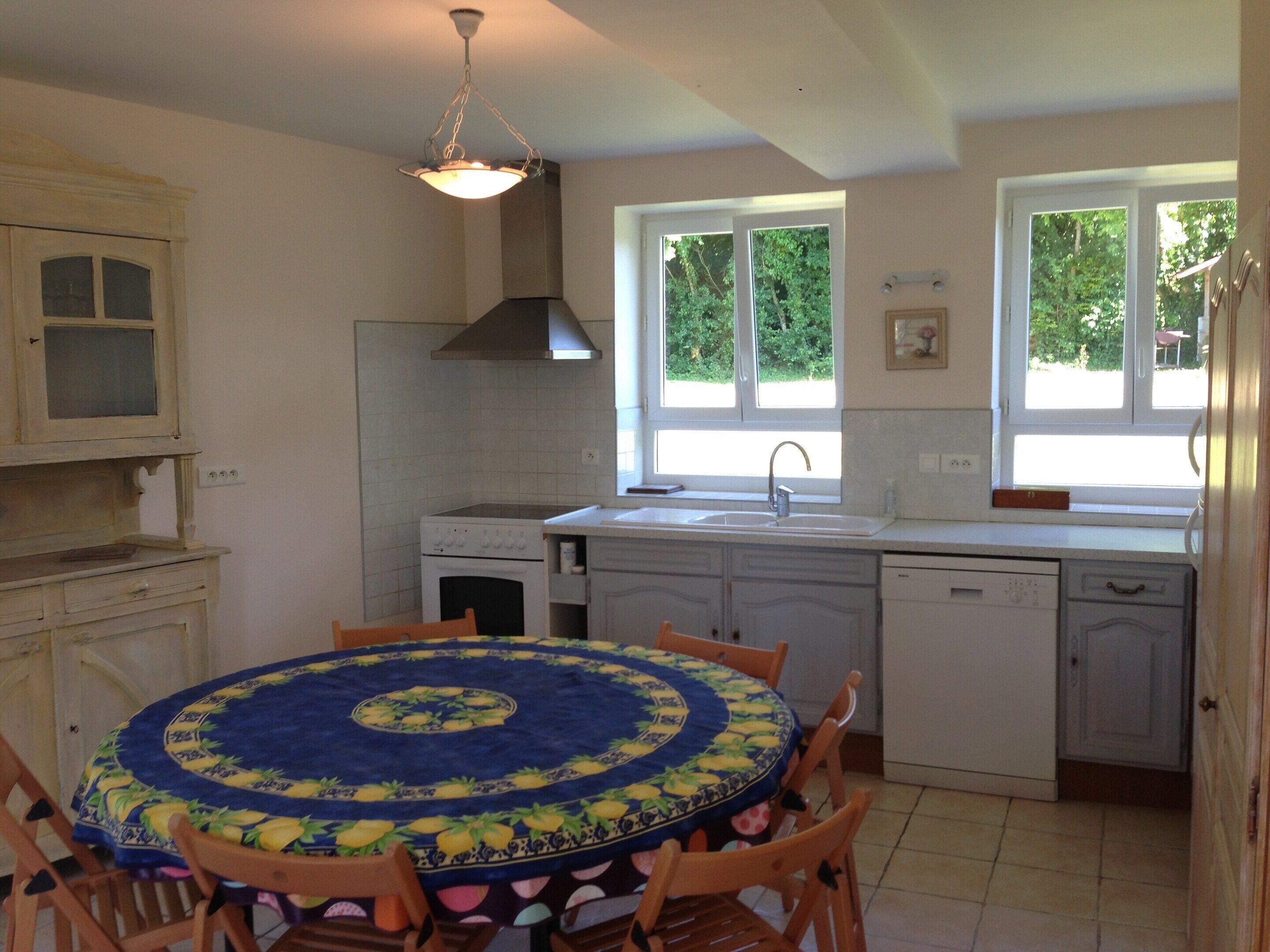 A kitchen with a round table, wooden chairs, a built-in oven, a sink, and a window with a view of greenery.