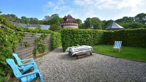 En uteplats med grusbeläggning, ett picknickbord och stolar, en häck och en byggnad med ett konformat tak.
