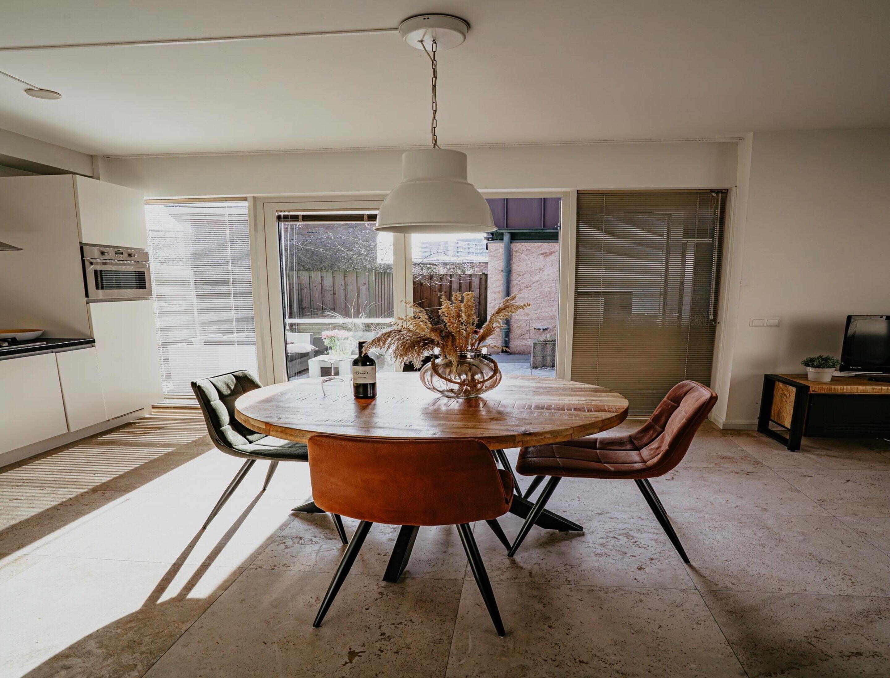 A dining area with a round table, chairs, a hanging light fixture, and a view of the outside through large windows.