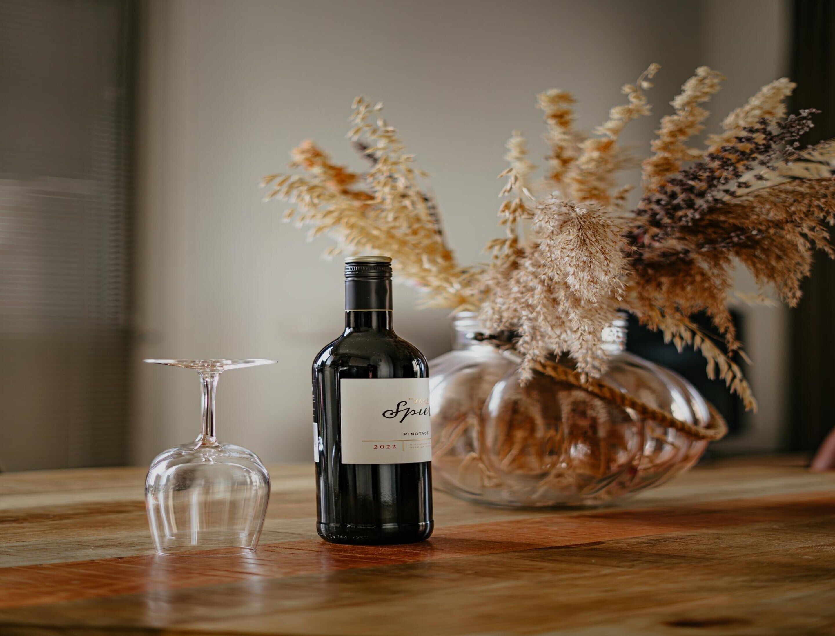 A wine bottle labeled 'Spel' and a glass on a wooden table, with dried flowers in a glass bowl in the background.