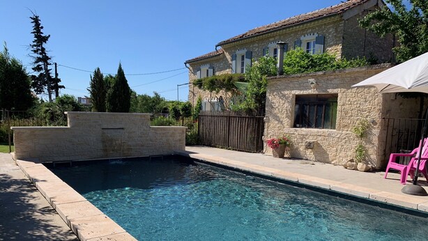 A swimming pool with a stone wall, a stone building with shutters, and a pink chair.