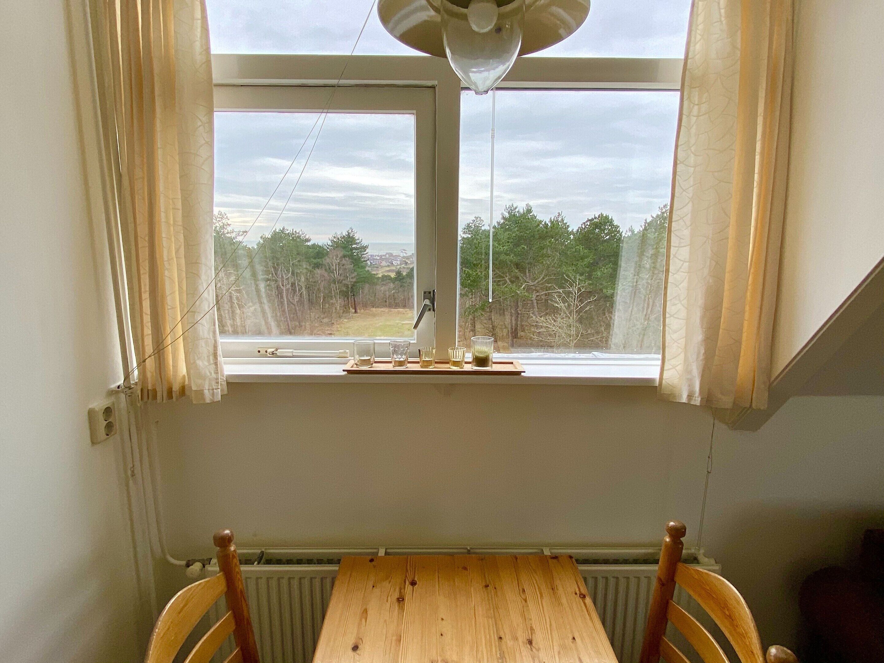 Ett rum med ett träbord och stolar, ett fönster med gardiner och utsikt över träd och en väg.