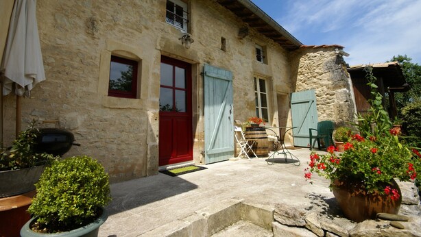 Un bâtiment en pierre avec une porte rouge, un volet bleu et un petit porche agrémenté de plantes en pot et de meubles d’extérieur.