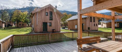 A wooden cabin with a deck, surrounded by a fenced area and other cabins, set in a mountainous landscape.