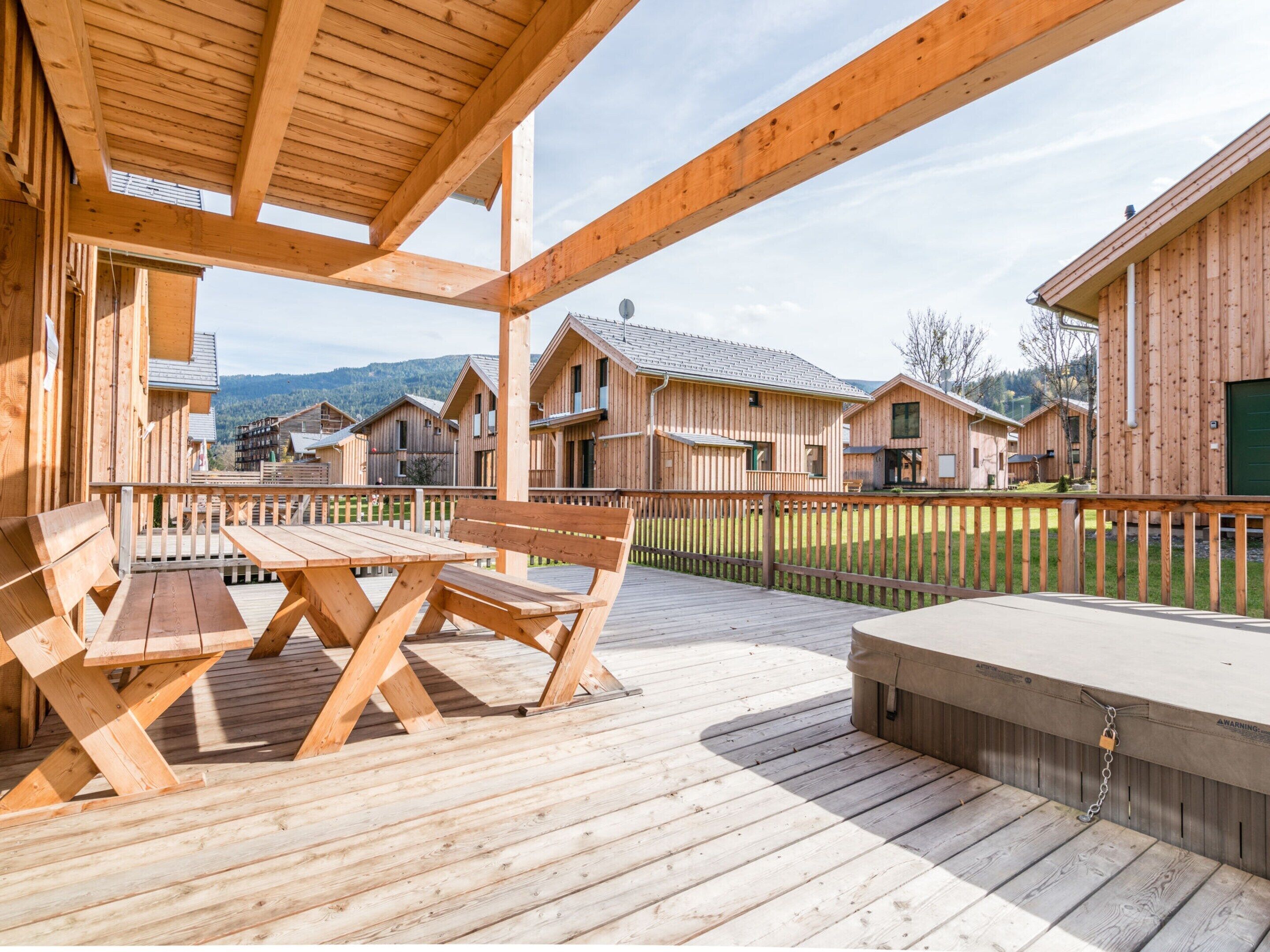 Una terraza de madera con una mesa y bancos, rodeada de cabañas de madera con vista a las montañas.