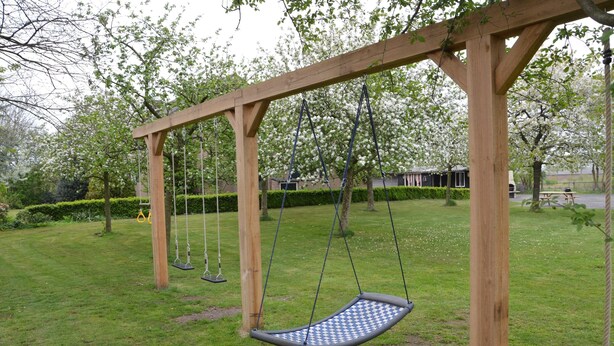 A wooden swing set in a park with a grassy area and trees. - Holiday Home in Elsendorp With Garden (Elsendorp)