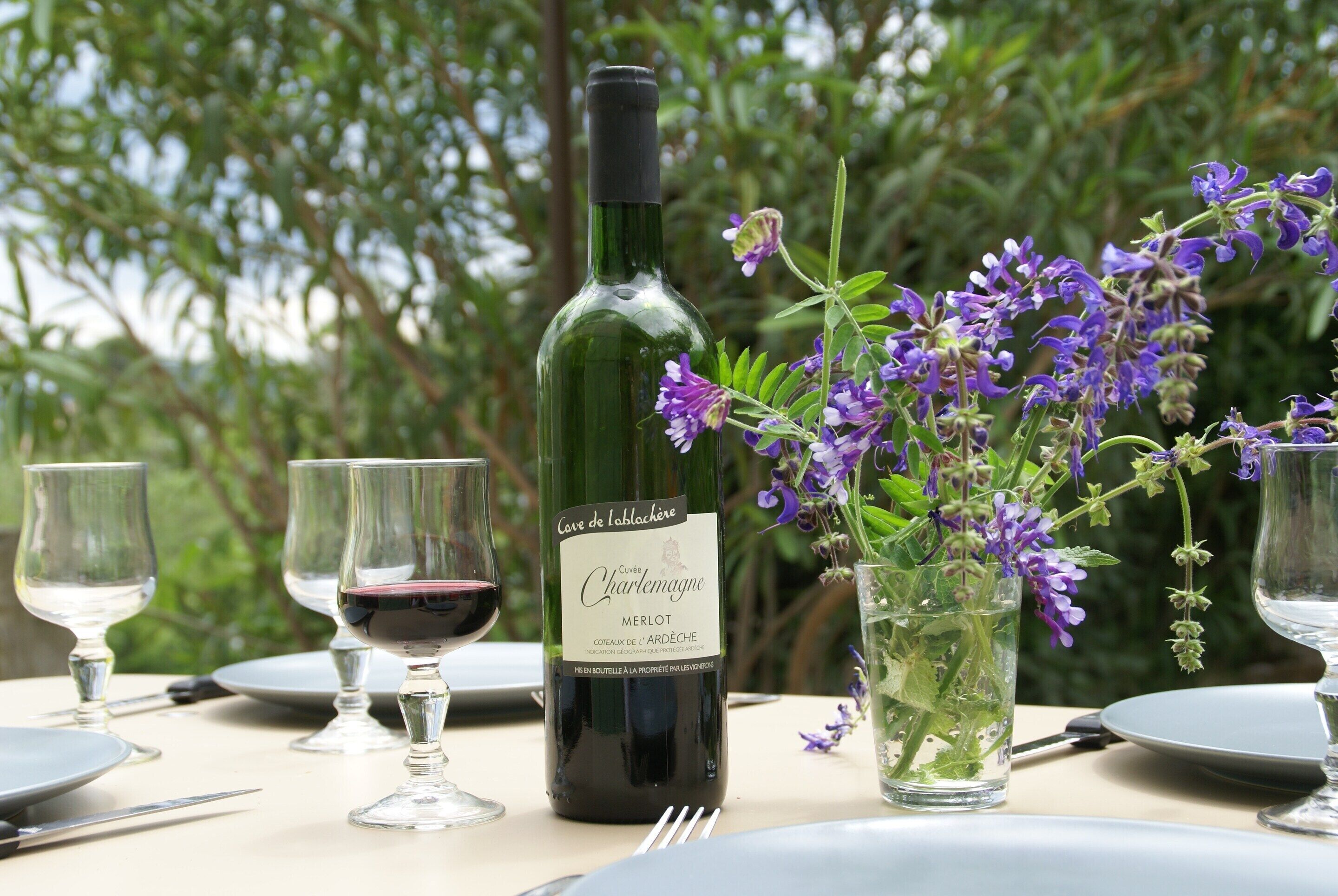 Mesa posta para refeição com garrafa de vinho, copos e um vaso com flores.