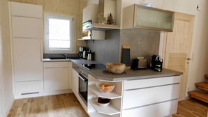 Uma cozinha compacta com armários brancos, forno embutido e micro-ondas. Há uma janela que oferece iluminação natural, e o piso é de madeira.