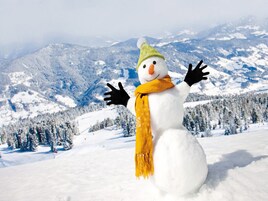 Un muñeco de nieve con sombrero verde y bufanda amarilla, de pie en un paisaje nevado con montañas al fondo.